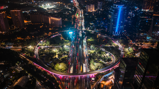 Aerial view of the Semanggi Interchange at night featuring illuminated circular highway ramps heavy traffic light trails and surrounding skyscrapers in Jakarta Indonesia.
