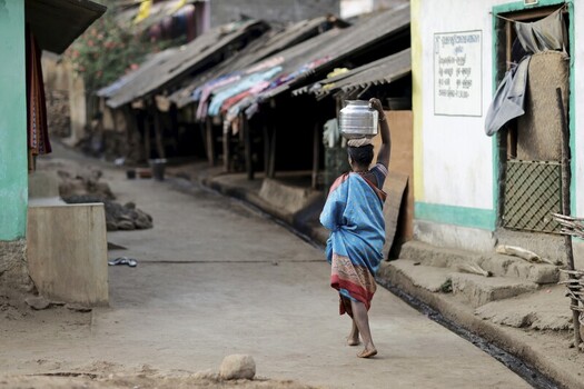 Odisha India 26 March 2026 View of a woman carrying water on her head along a dusty street lined with weathered homes a scene of daily life. Odisha India 26 March 2026 View of a woman carrying water on her head along a dusty street lined with weathered homes a scene of daily life.
