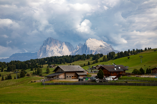 View of traditional wooden mountain huts nestled in a lush green meadow with the dramatic peaks of the Dolomites rising under a cloudy sky in Rocca Pietore Veneto Italy. View of traditional wooden mountain huts nestled in a lush green meadow with the dramatic peaks of the Dolomites rising under a cloudy sky in Rocca Pietore Veneto Italy.