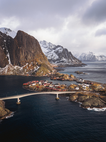 Aerial view of red cabins cluster tightly on islands linked by a bridge against a backdrop of snow dusted mountains and moody skies Sakris&oslash;ya Nordland Norway.