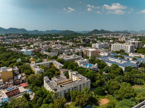 Aerial view of the cityscape of Udaipur featuring dense urban architecture lush green trees and distant hills under a clear blue sky in Udaipur Rajasthan India.