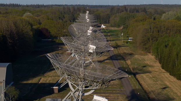 Aerial view of the Westerbork Synthesis Radio Telescope dishes aligned in a row surrounded by lush green forests and distant wind turbines in Westerbork Drenthe Netherlands. Aerial view of the Westerbork Synthesis Radio Telescope dishes aligned in a row surrounded by lush green forests and distant wind turbines in Westerbork Drenthe Netherlands.
