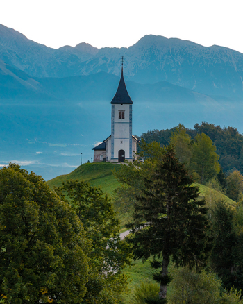 Aerial view of the Church of Saints Primus and Felician perched on a grassy hilltop with the Julian Alps in the background in Jamnik Kranj Slovenia.