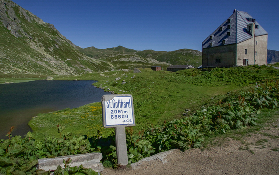 View of Sankt Gotthard Pass featuring a historic stone building a mountain lake and a signpost under a clear blue sky Airolo Ticino Switzerland.