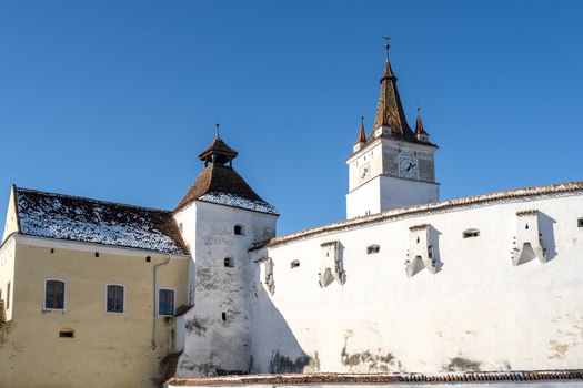 View of the historic Prejmer Fortified Church with its white defensive walls clock tower and snow dusted roofs under a clear blue sky in Prejmer Brasov Romania. View of the historic Prejmer Fortified Church with its white defensive walls clock tower and snow dusted roofs under a clear blue sky in Prejmer Brasov Romania.