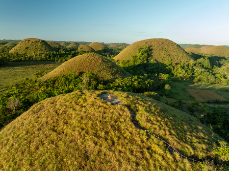 Aerial view of the Chocolate Hills with a person standing on a grassy peak surrounded by iconic conical hills and lush tropical vegetation in Batuan Central Visayas Philippines. Aerial view of the Chocolate Hills with a person standing on a grassy peak surrounded by iconic conical hills and lush tropical vegetation in Batuan Central Visayas Philippines.