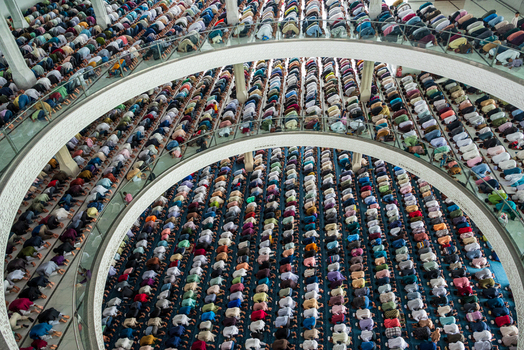 Dhaka Bangladesh 19 June 2026 Aerial view of a large crowd of men praying in rows inside a mosque with curved balconies and patterned carpets.