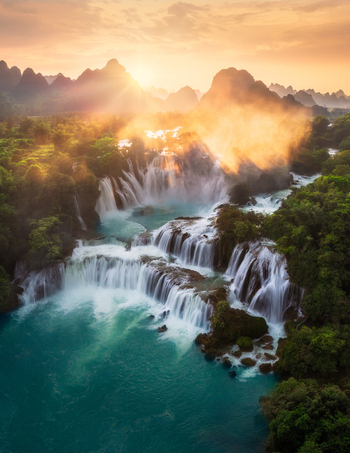 Aerial view of the majestic Ban Gioc Waterfall cascading over limestone cliffs and lush greenery under the warm glow of the setting sun Ban Gioc Waterfall Cao Bang Vietnam.