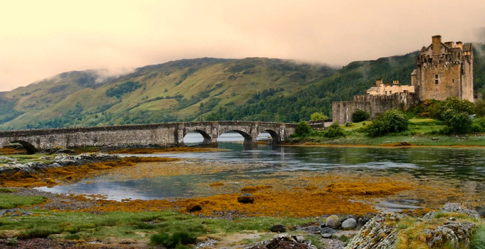 Aerial view of Eilean Donan Castle with its historic stone bridge spanning the water surrounded by lush green hills under a soft golden hour sky in Dornie Scotland United Kingdom.