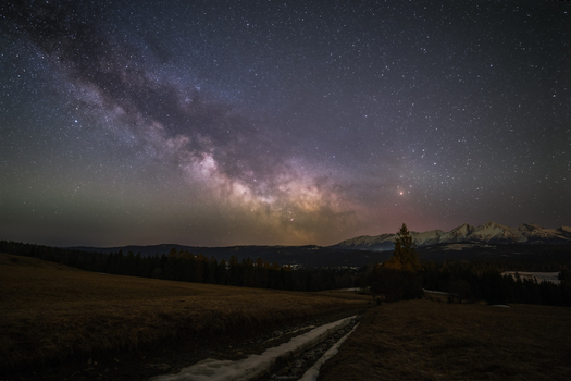 View of the Milky Way galaxy stretching over the dark silhouette of the Tatra Mountains and a grassy field with a path in Poland. View of the Milky Way galaxy stretching over the dark silhouette of the Tatra Mountains and a grassy field with a path in Poland.
