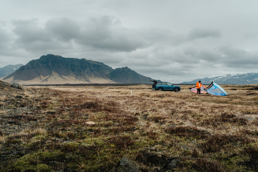 View of a person in an orange jacket preparing a kite on the mossy volcanic plains with mountains in the background in Borgarbyggd Iceland. View of a person in an orange jacket preparing a kite on the mossy volcanic plains with mountains in the background in Borgarbyggd Iceland.