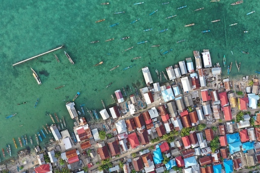 Aerial view of Pulau Maginti featuring stilt houses over the turquoise sea with many small wooden fishing boats moored along the shoreline Maginti South East Sulawesi Indonesia.