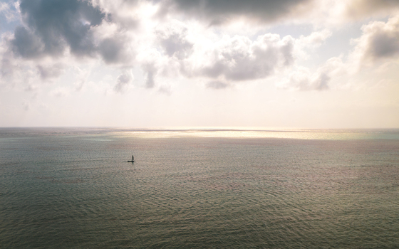 Aerial view of a lone sailboat cuts through the vast expanse of the ocean under a sky of soft muted tones Paje Unguja South Region Tanzania. Aerial view of a lone sailboat cuts through the vast expanse of the ocean under a sky of soft muted tones Paje Unguja South Region Tanzania.