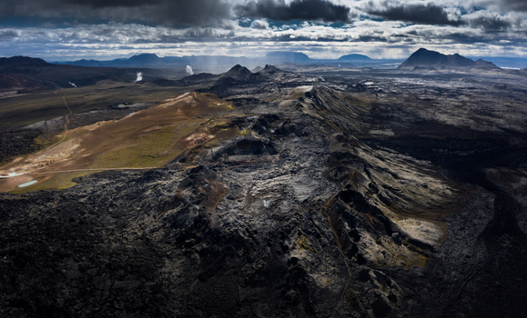 Aerial view of a vast volcanic landscape with dark lava fields rugged craters and geothermal steam rising under a cloudy sky in Iceland.