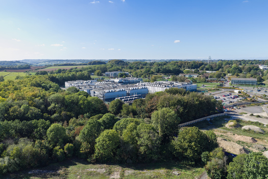 Aerial view of a large data center facility surrounded by dense green forest and trees under a clear blue sky United Kingdom.