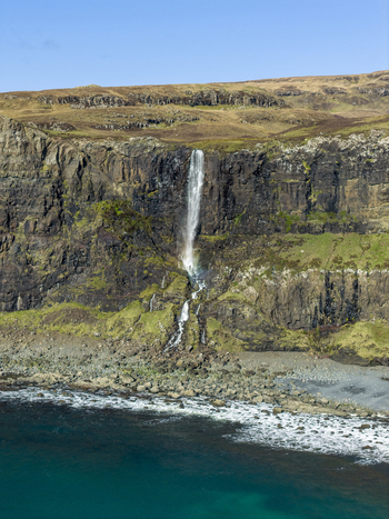 Aerial view of Talisker Bay Beach waterfall cascading down rugged cliffs into the turquoise sea under a clear blue sky in Isle of Skye Scotland United Kingdom. Aerial view of Talisker Bay Beach waterfall cascading down rugged cliffs into the turquoise sea under a clear blue sky in Isle of Skye Scotland United Kingdom.