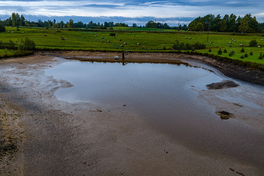 Aerial view of a small pond surrounded by green fields with hay bales and distant trees under an overcast sky in Latvia.