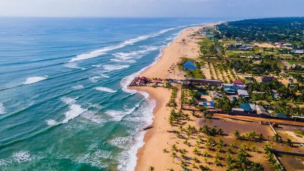 Aerial view of a sandy coastline with turquoise waves crashing on the shore palm trees and coastal buildings in Ghana. Aerial view of a sandy coastline with turquoise waves crashing on the shore palm trees and coastal buildings in Ghana.