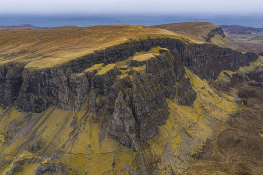 Aerial view of The Quiraing featuring dramatic rock formations steep cliffs and golden grassy plateaus under a misty sky Isle of Skye Scotland United Kingdom. Aerial view of The Quiraing featuring dramatic rock formations steep cliffs and golden grassy plateaus under a misty sky Isle of Skye Scotland United Kingdom.
