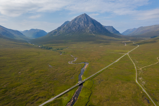 Aerial view of Buachaille Etive Mor mountain peak rising above the green landscape of Glen Coe with a winding river and a road crossing a bridge in Ballachulish Scotland United Kingdom. Aerial view of Buachaille Etive Mor mountain peak rising above the green landscape of Glen Coe with a winding river and a road crossing a bridge in Ballachulish Scotland United Kingdom.