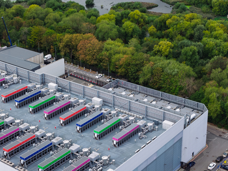 Aerial view of a data center with rows of cooling units on the roof adjacent to a wooded area and parking lot in United Kingdom.