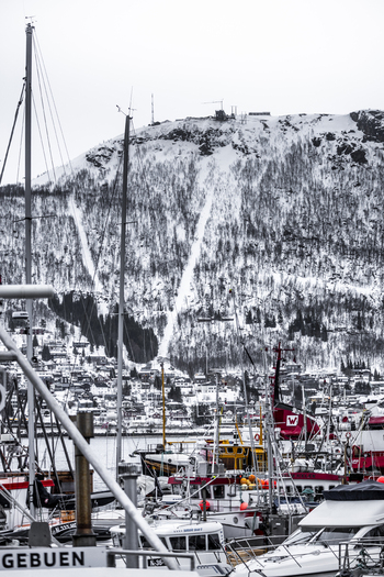 View of a busy harbor filled with fishing boats and yachts against the backdrop of a snow covered mountain and hillside houses in Tromso Troms Norway. View of a busy harbor filled with fishing boats and yachts against the backdrop of a snow covered mountain and hillside houses in Tromso Troms Norway.