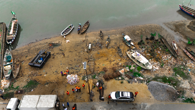 Aerial view of a riverbank with numerous small wooden boats people walking near piles of debris and a parked car on the shore in Minima Rivers Nigeria.