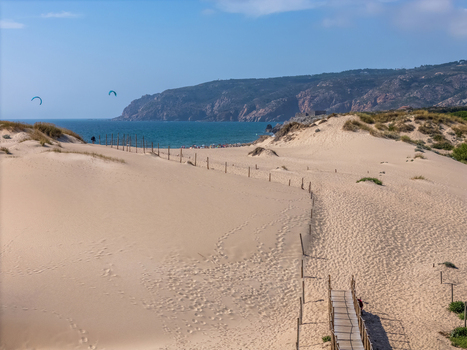 Aerial view of Praia do Guincho with expansive sand dunes a wooden walkway leading towards the ocean and kite surfers in the distance under a clear blue sky in Cascais Lisbon Portugal. Aerial view of Praia do Guincho with expansive sand dunes a wooden walkway leading towards the ocean and kite surfers in the distance under a clear blue sky in Cascais Lisbon Portugal.