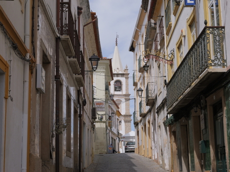 View of the historic Se de Portalegre cathedral tower rising above a narrow cobblestone street lined with traditional buildings and balconies in Portalegre Portalegre District Portugal.