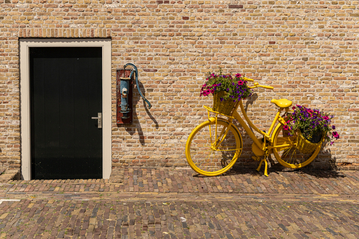 View of a yellow bicycle with flower baskets parked against a brick wall next to a black door and an old water pump in Heusden North Brabant Netherlands.
