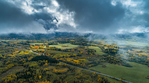 Aerial view of a winding road through a vibrant autumn forest and green fields under a dramatic cloudy sky in Latvia. Aerial view of a winding road through a vibrant autumn forest and green fields under a dramatic cloudy sky in Latvia.