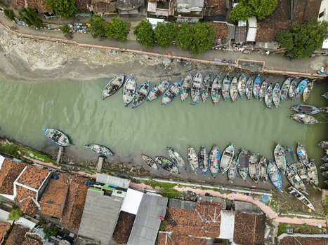 Aerial view of boats docked along the river contrasting with the red tiled rooftops of buildings casting shadows in the afternoon sun Jepara Central Java Indonesia.