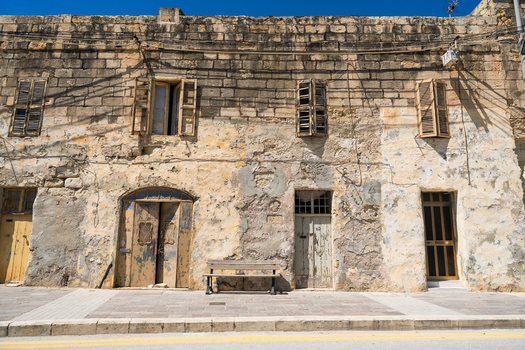 View of a traditional weathered limestone building facade with old wooden shutters and doors under a clear blue sky in Marsaskala Southern Region Malta. View of a traditional weathered limestone building facade with old wooden shutters and doors under a clear blue sky in Marsaskala Southern Region Malta.