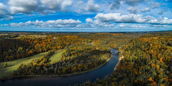Aerial view of the Gauja River winding through a dense forest with vibrant autumn foliage and a bridge under a blue sky in Sigulda Sigulda Municipality Latvia. Aerial view of the Gauja River winding through a dense forest with vibrant autumn foliage and a bridge under a blue sky in Sigulda Sigulda Municipality Latvia.