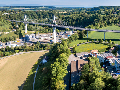 Aerial view of Poya Bridge spanning a lush green valley with industrial structures and rolling hills under a clear sky Fribourg Fribourg Switzerland.