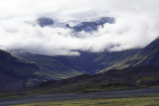 View of Skaftafell mountains with snow capped peaks partially obscured by low hanging clouds above mossy green slopes and dark volcanic plains in Skaftafell Iceland. View of Skaftafell mountains with snow capped peaks partially obscured by low hanging clouds above mossy green slopes and dark volcanic plains in Skaftafell Iceland.