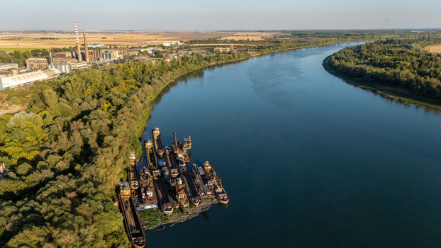 Aerial view of a group of moored barges on the Sava River near a forested bank with an industrial facility in the background in Sremska Mitrovica Vojvodina Serbia.