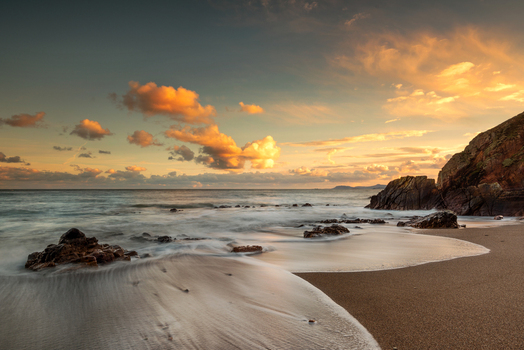 View of a sandy beach at sunset with waves washing over rocks and golden clouds in the sky near Howth Ireland. View of a sandy beach at sunset with waves washing over rocks and golden clouds in the sky near Howth Ireland.