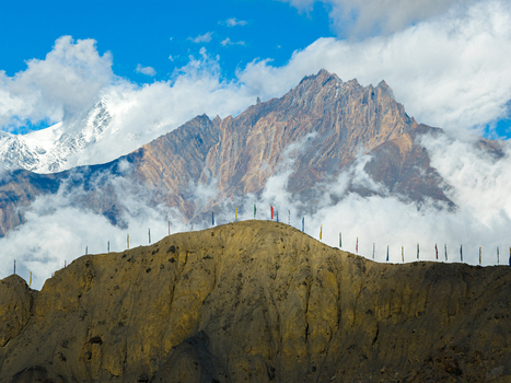 Aerial view of a rocky mountain ridge adorned with colorful prayer flags set against a backdrop of towering snow capped Himalayan peaks and drifting clouds in Nepal.