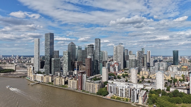 London United Kingdom 16 June 2026 Aerial view of the Canary Wharf skyline featuring modern skyscrapers along the River Thames under a cloudy blue sky.