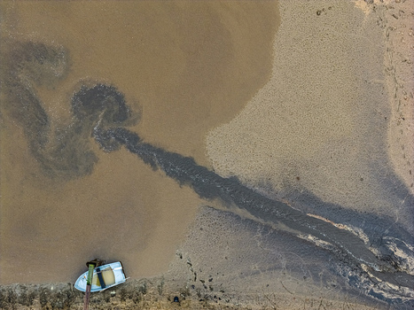 Aerial view of a small boat moored on a sandy shoreline near a dark plume of sediment flowing into murky water in Latvia.