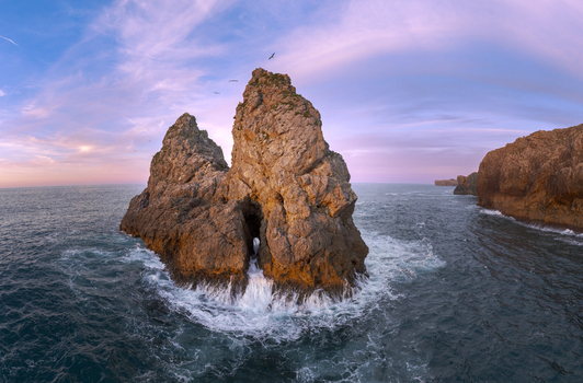 Aerial view of a dramatic sea stack rising from the ocean with waves crashing against its base under a vibrant pink and blue sunset sky in Nueva Asturias Spain.