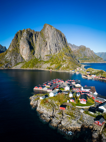 Aerial view of Hamnoy village with red rorbuer cabins on a rocky island surrounded by deep blue water and dramatic mountains under a clear sky in Hamnoy Nordland Norway. Aerial view of Hamnoy village with red rorbuer cabins on a rocky island surrounded by deep blue water and dramatic mountains under a clear sky in Hamnoy Nordland Norway.