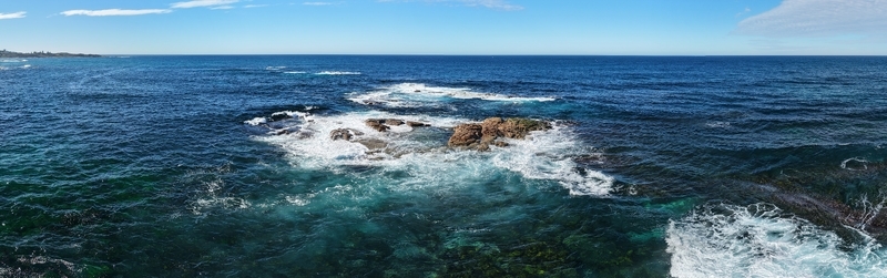 Aerial view of the rugged coastline where frothy white waves crash against the dark rocks under a clear blue sky Kiama New South Wales Australia. Aerial view of the rugged coastline where frothy white waves crash against the dark rocks under a clear blue sky Kiama New South Wales Australia.