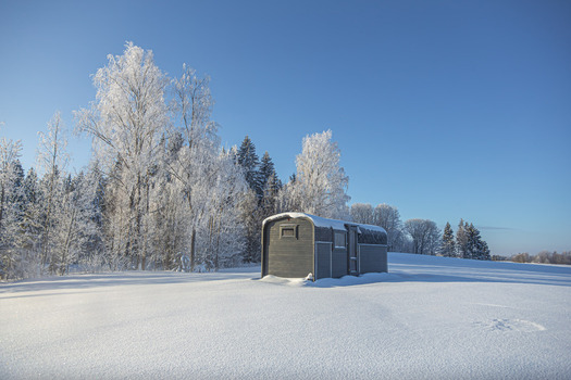 View of a small dark wooden cabin situated in a vast snow covered field surrounded by frost covered trees under a clear blue sky in Latvia. View of a small dark wooden cabin situated in a vast snow covered field surrounded by frost covered trees under a clear blue sky in Latvia.