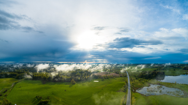 Aerial view of a rural landscape with green fields a winding road and a small lake under a dramatic sky with low hanging clouds in Latvia. Aerial view of a rural landscape with green fields a winding road and a small lake under a dramatic sky with low hanging clouds in Latvia.