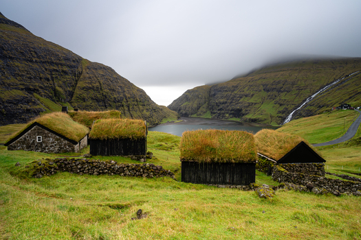 View of traditional turf roofed houses nestled in a lush green valley with a mountain lake and a waterfall in the background under a cloudy sky in the Faroe Islands.