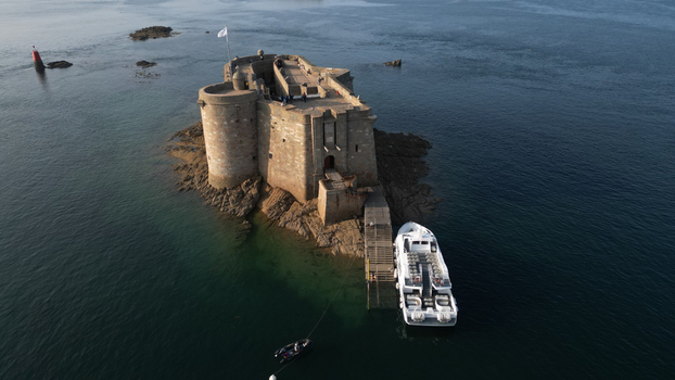 Aerial view of the imposing Ch&acirc;teau du Taureau standing resiliently against the dark rippling sea a stark contrast to the bright boat docked nearby Plouezoc h Bretagne France.