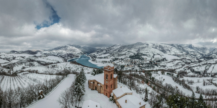 Aerial view of a historic brick church surrounded by snow covered hills trees and a lake in the background under a cloudy winter sky in Fiastra Marche Italy. Aerial view of a historic brick church surrounded by snow covered hills trees and a lake in the background under a cloudy winter sky in Fiastra Marche Italy.
