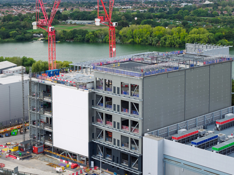 Aerial view of a large data center under construction with steel framework and red cranes situated near a lake in the United Kingdom.